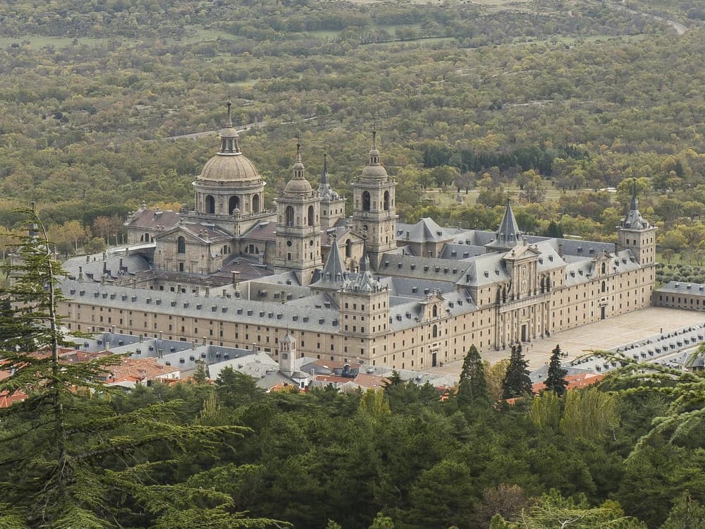 The austere grey granite facade of El Escorial with the Sierra de Guadarrama mountains rising behind under a dramatic sky