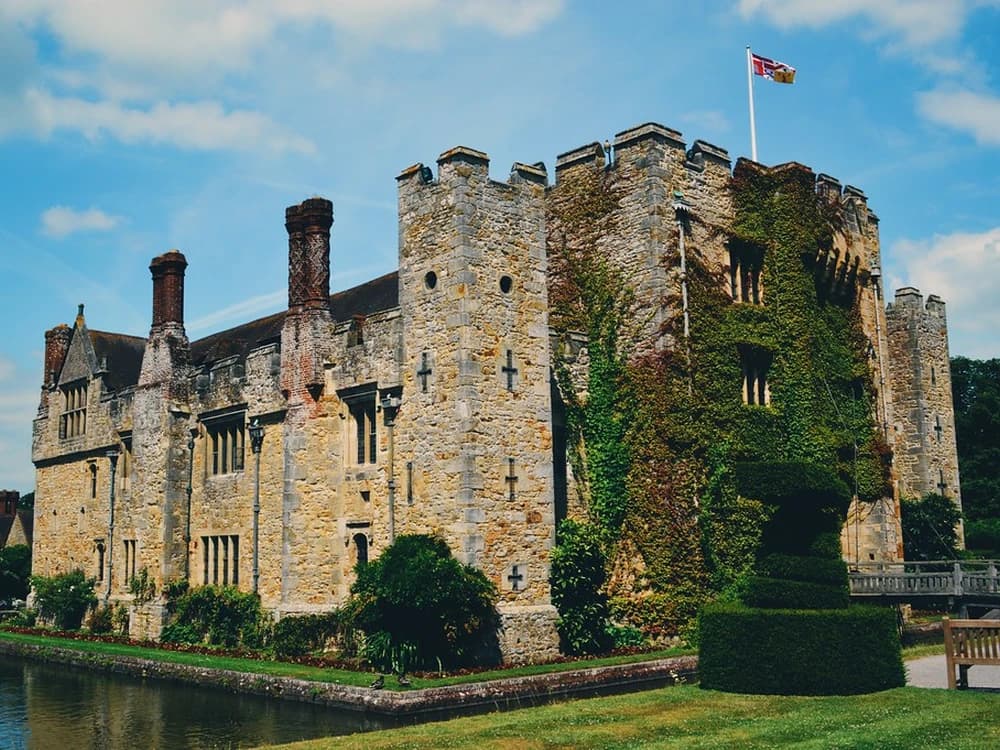 Hever Castle's medieval gatehouse and stone towers reflected in its double moat on a still autumn morning, surrounded by trees in russet and gold
