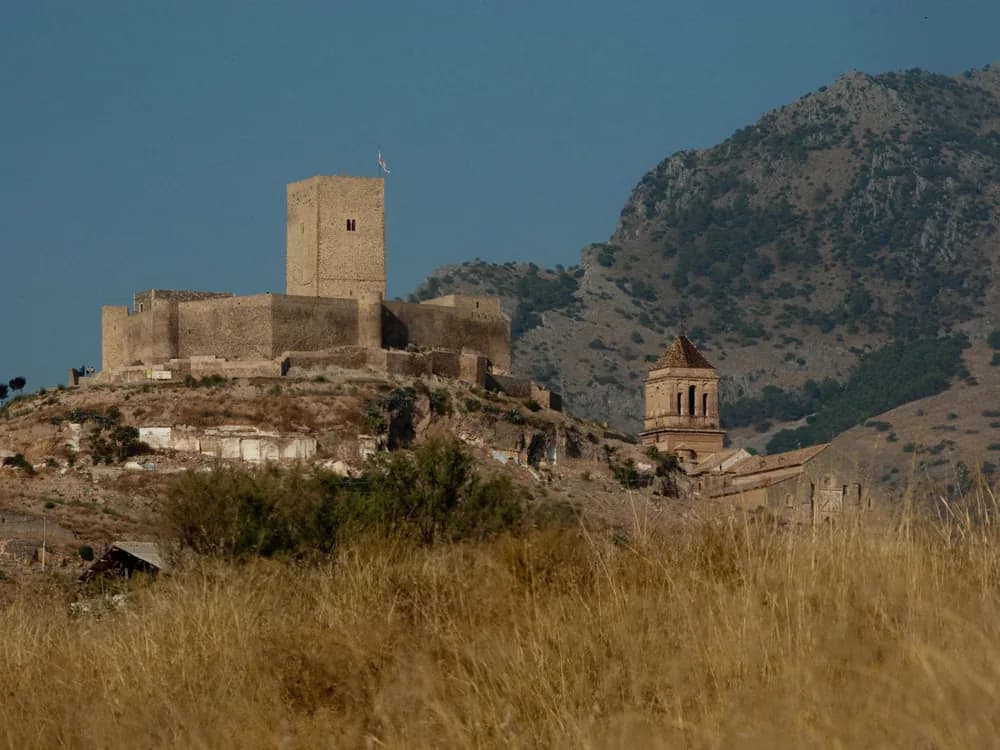 Castle of Alcaudete on its limestone hilltop above the olive groves of Jaén province — the frontier fortress of the Order of Calatrava, with the Torre del Homenaje and defensive walls spanning Caliphate, Almohad, and Christian military-order periods