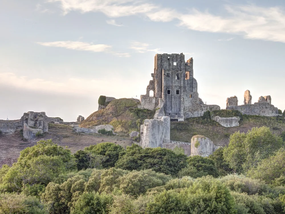 Corfe Castle ruins on the chalk hilltop above the Isle of Purbeck — the deliberately slighted medieval fortress with its leaning towers and broken walls, Dorset, England