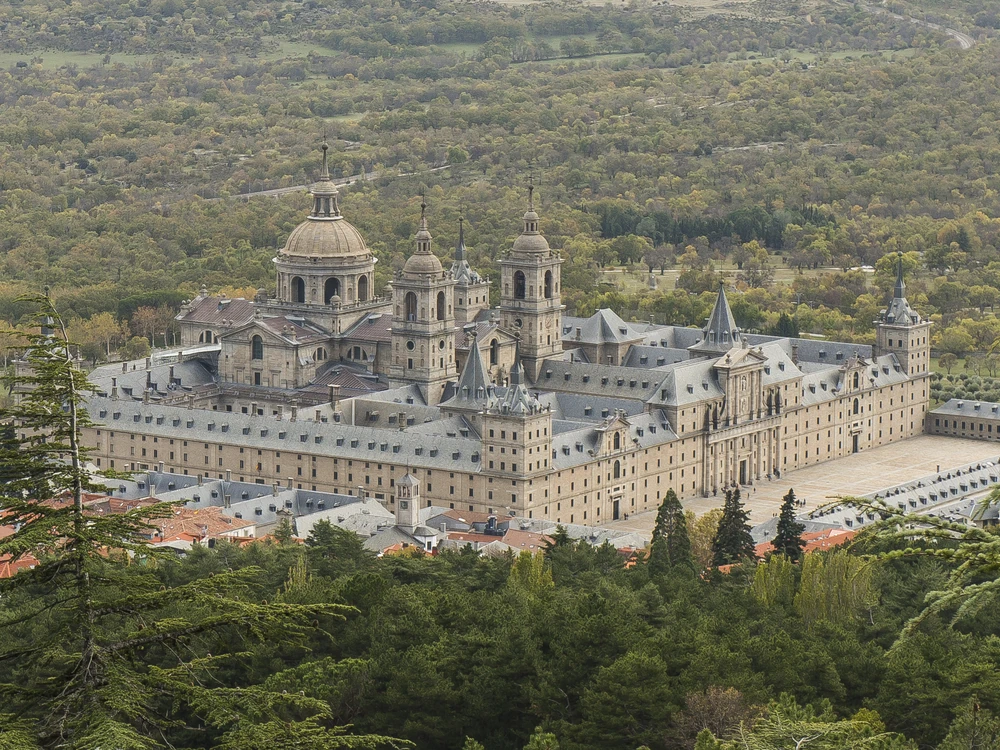 The austere grey granite facade of El Escorial with the Sierra de Guadarrama mountains rising behind under a dramatic sky