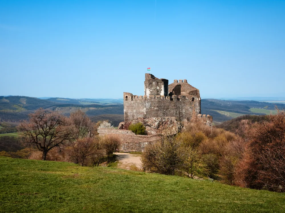 Hollókő Castle on its basalt ridge above the UNESCO World Heritage village in Nógrád County, Hungary — the 13th–14th-century ruins overlooking the traditional Palóc wooden-porched houses of the village below