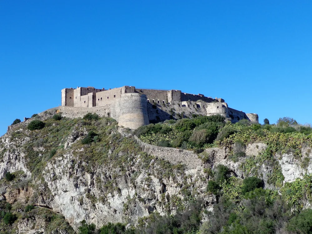 Milazzo Castle's multi-period fortification rising above the Borgo Antico on the Milazzo peninsula, with the Tyrrhenian Sea and the distant Aeolian Islands visible beyond