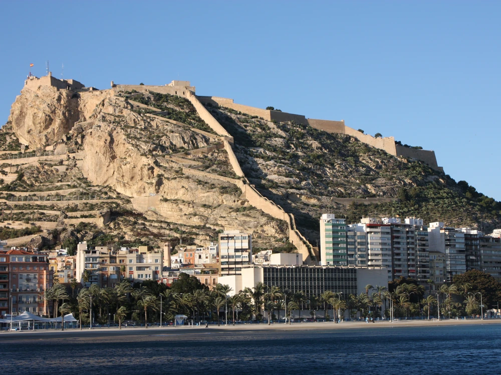 Santa Bárbara Castle's medieval walls and towers rising above Alicante city and the Mediterranean coast, with Postiguet beach glinting in the afternoon sun below