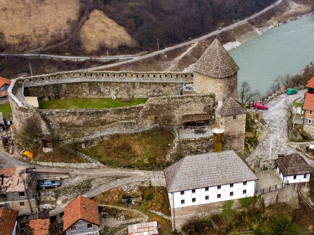 Vranduk Fortress rising above the Bosna River gorge in central Bosnia — the medieval royal gateway fortress on its limestone rock spur, with the river channeled below the walls