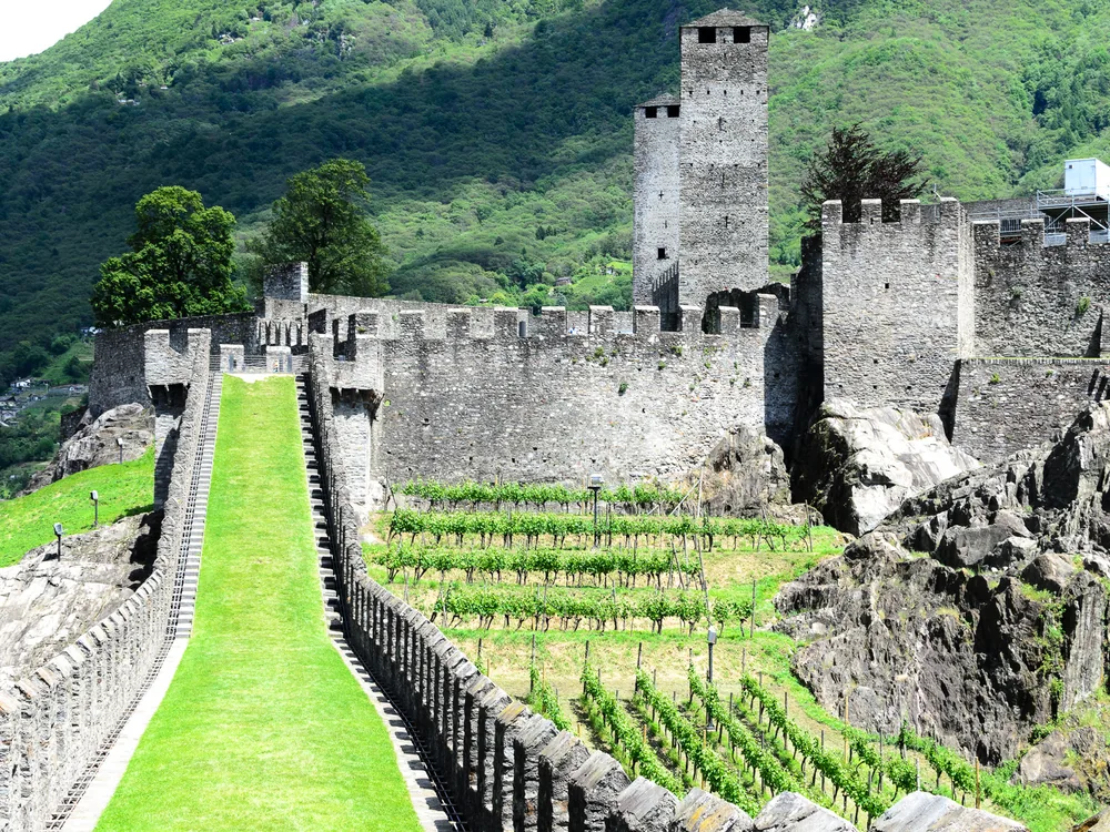 The three UNESCO World Heritage castles of Bellinzona, Switzerland — Castelgrande in the foreground with its twin towers, Montebello on the hill above, and Sasso Corbaro on the highest spur, connected by the Murata fortification walls